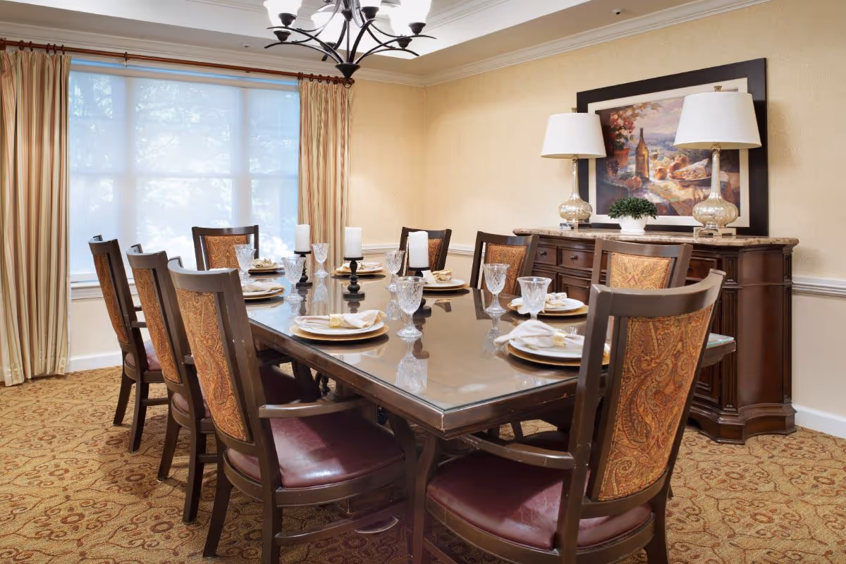A formal dining room with a large rectangular glass-top table set for eight people. The table is surrounded by wooden chairs with patterned upholstery. The table is set with plates, napkins, and crystal glasses, and there are three candle holders with white candles in the center. The room has beige walls, a patterned carpet, and a large window with sheer white curtains and gold drapes. Against the wall is a wooden sideboard with two matching lamps and a framed painting above it.