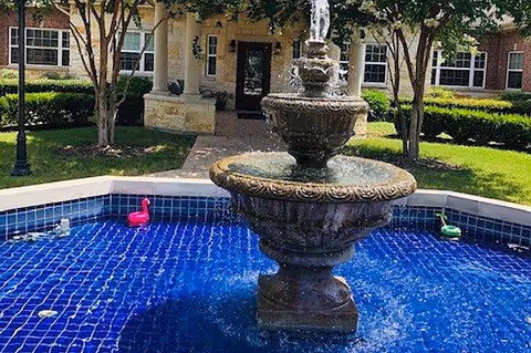 Ornate three-tier fountain in a blue-tiled courtyard pool with a residential building entrance and landscaping in the background.