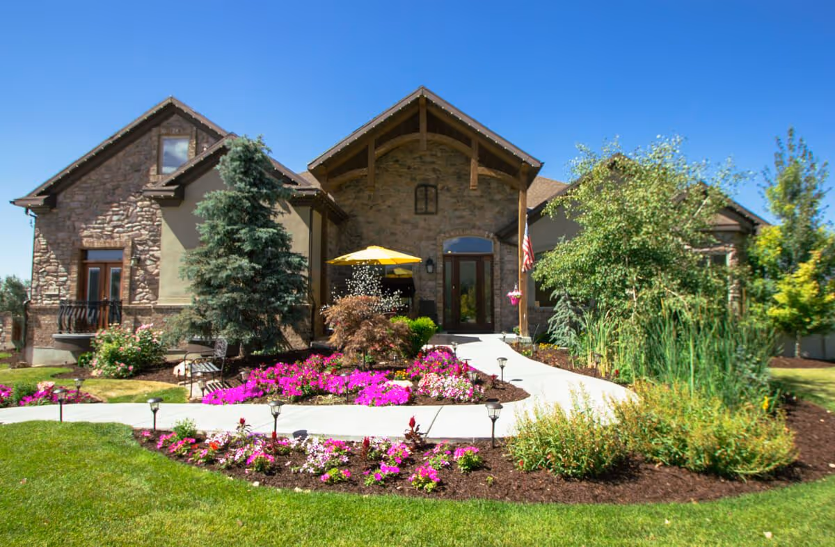 Stone-front residential building entrance with a curved walkway, colorful flower beds, shrubs, and a yellow umbrella under a clear blue sky.
