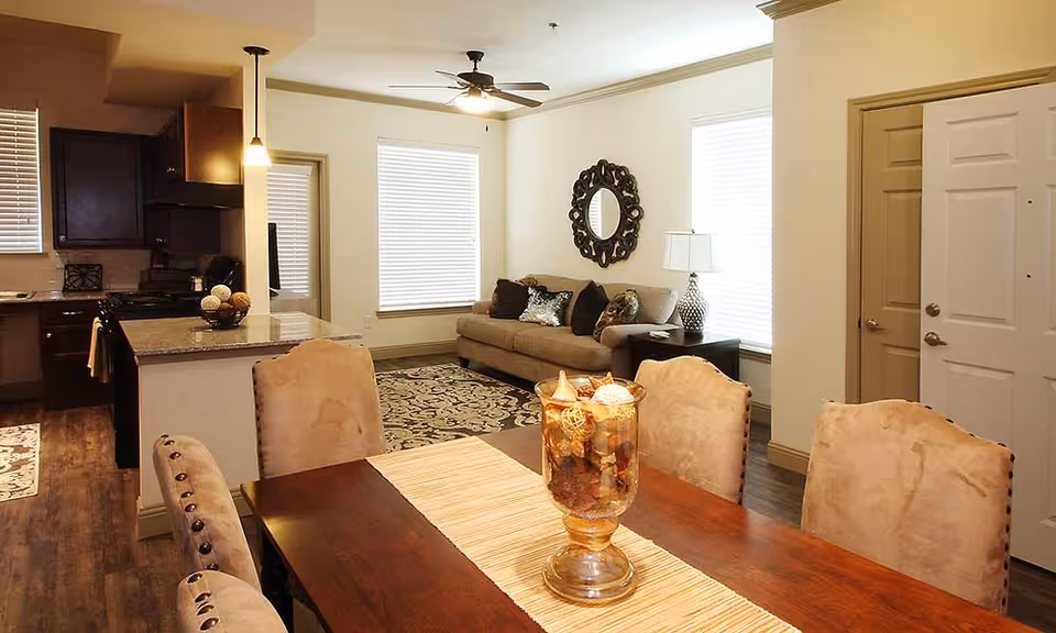 Open-plan dining area with a wooden table and upholstered chairs, a living room with a sofa and decorative mirror, and a kitchen visible in the background.
