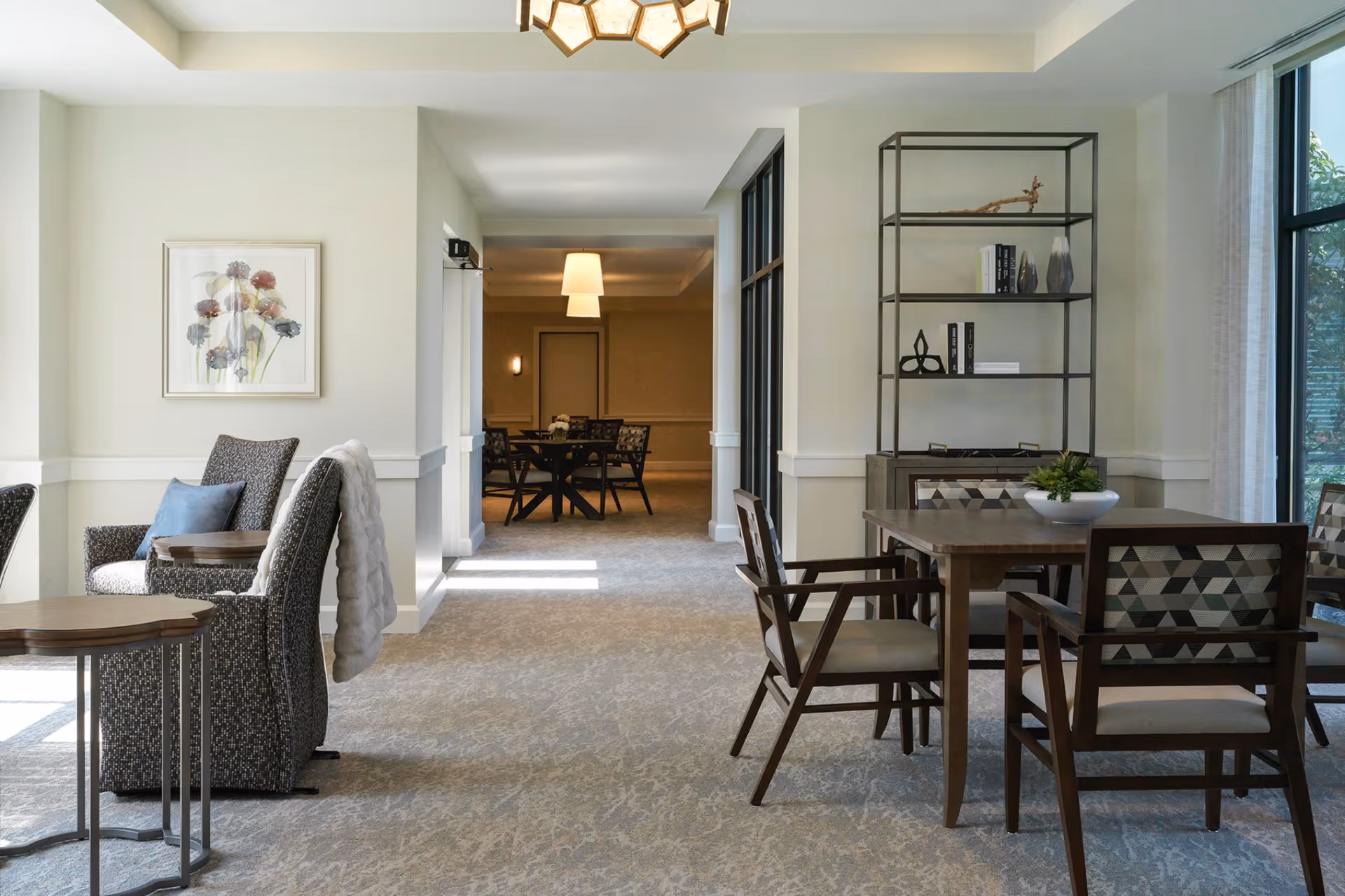 A bright and spacious common area in a senior living facility featuring comfortable armchairs with a small side table on the left, a wooden dining table with four chairs on the right, and a decorative shelving unit with books and ornaments. Large windows allow natural light to fill the room, and a hallway with additional seating and lighting fixtures is visible in the background.