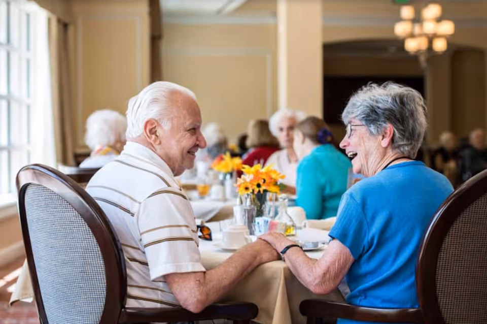 Two elderly people sitting at a dining table in a well-lit room, holding hands and smiling at each other. Other elderly individuals are seated at tables in the background, with flowers and dining items on the tables.