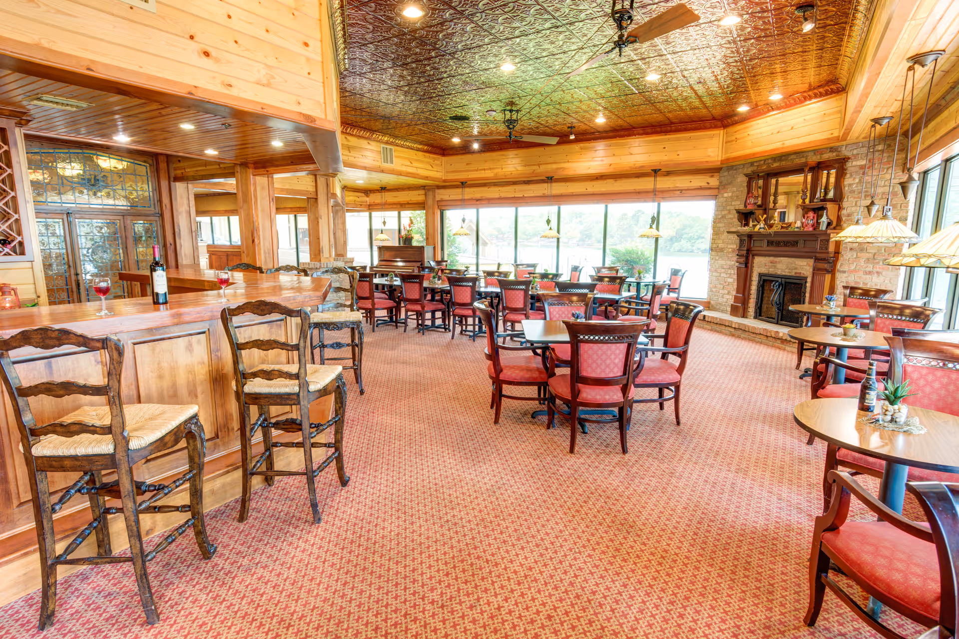 A spacious dining room with multiple round tables and red cushioned chairs arranged neatly. There is a wooden bar with high chairs on the left side, a fireplace with a wooden mantle on the right, and large windows providing a view of greenery outside. The ceiling has a decorative pattern with ceiling fans and recessed lighting.