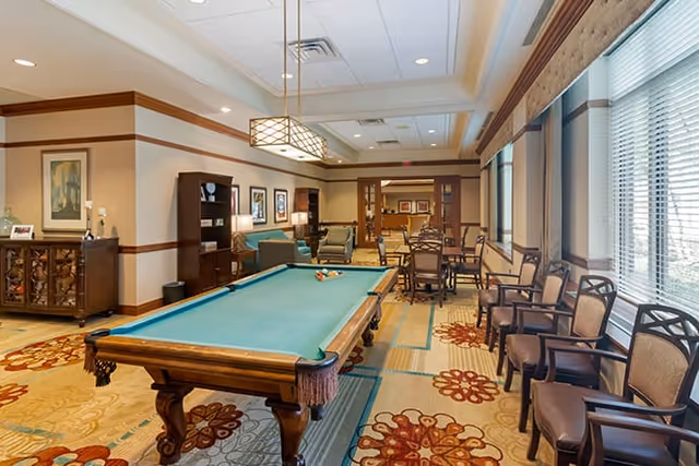 Well-lit common room featuring a pool table, multiple seating areas, dining tables, and large windows.