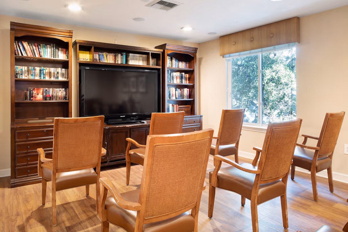 A cozy common living room with six wooden armchairs facing a large flat-screen TV set in a built-in bookshelf and a window letting in daylight.