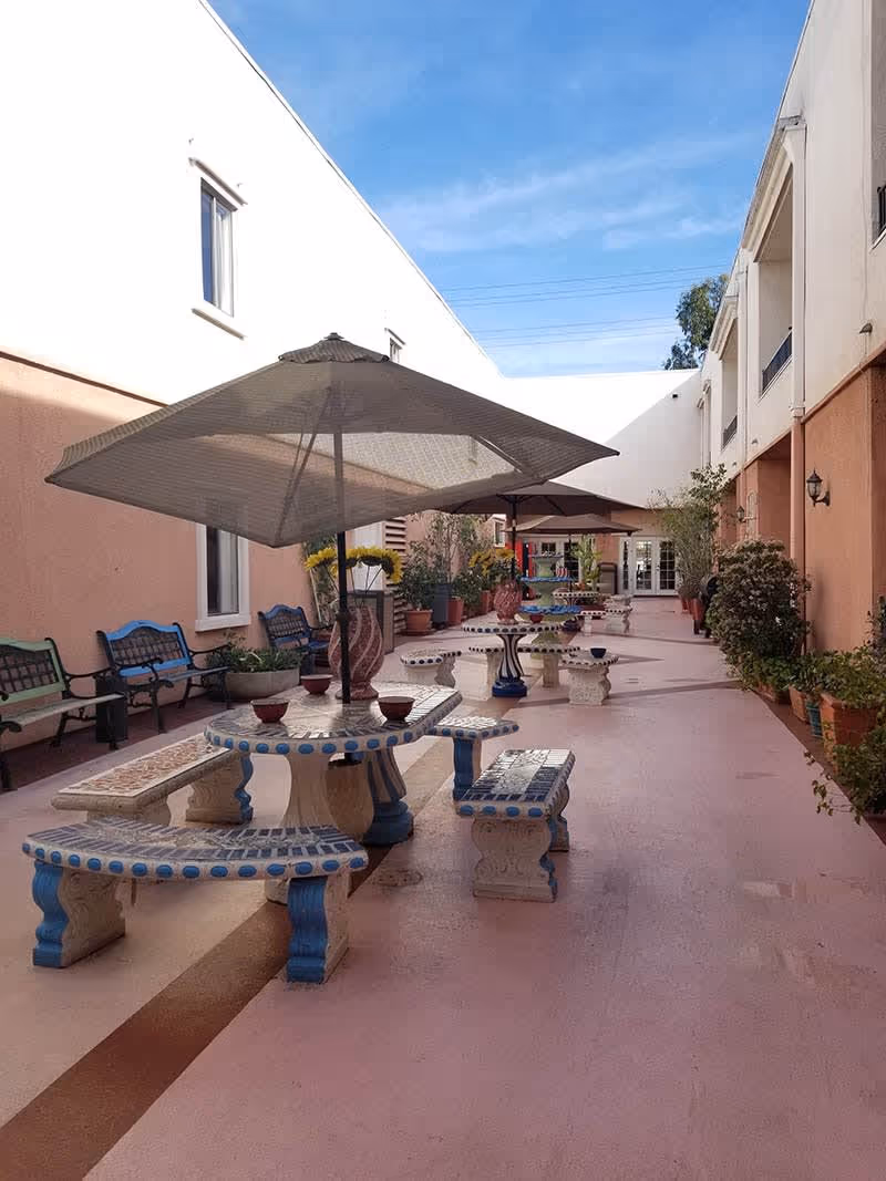 Outdoor courtyard area at Fine Gold Manor with stone tables and benches featuring blue accents, large umbrellas providing shade, potted plants along the walls, and benches against the building walls under a clear blue sky.