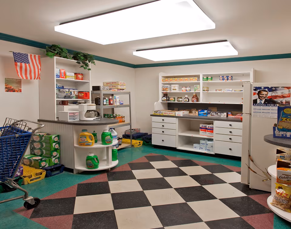 Small convenience store area inside a senior living facility with shelves of snacks and cleaning supplies, a shopping cart, and a checkout counter.