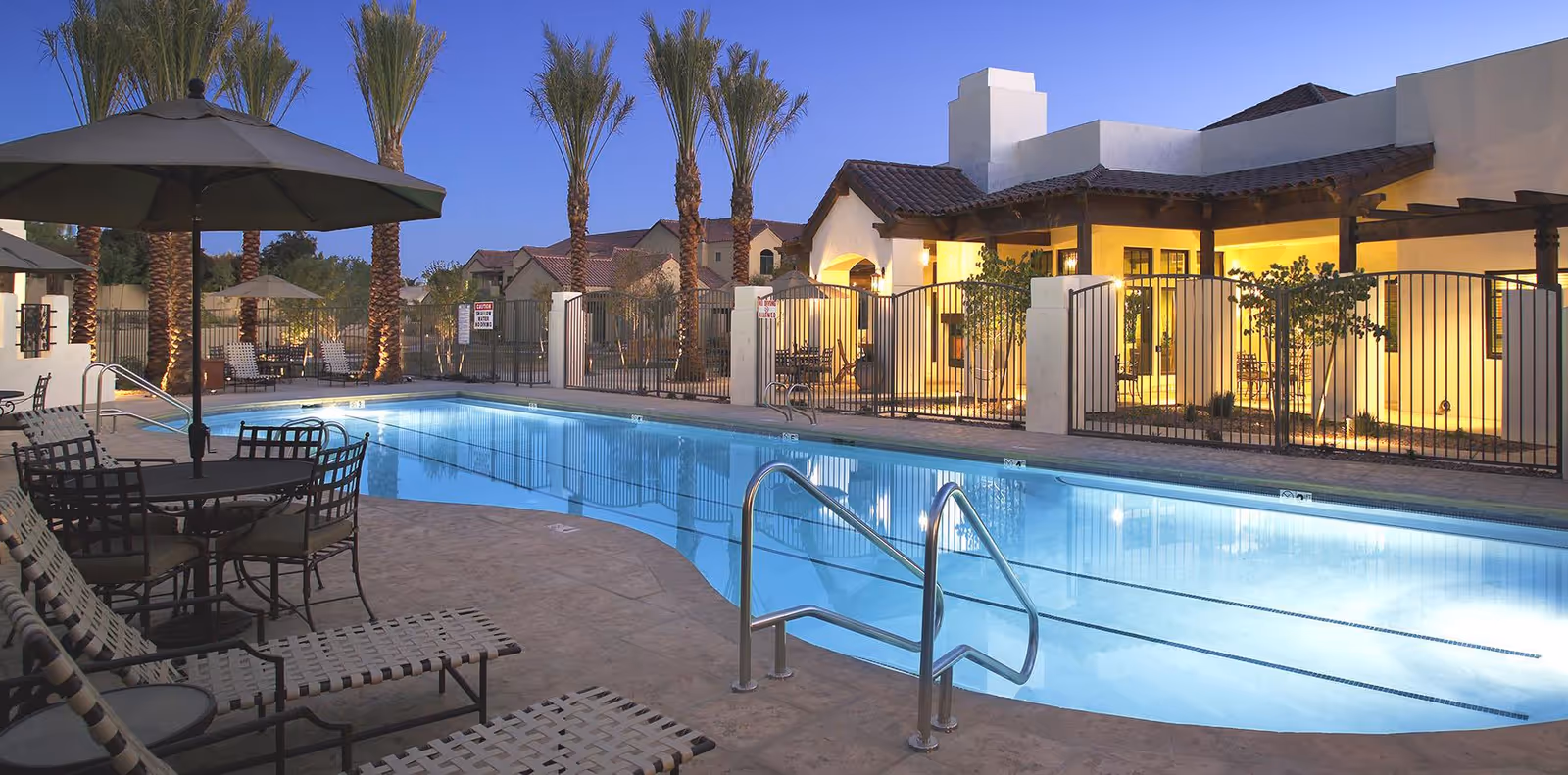 Outdoor swimming pool area at dusk with lounge chairs, tables with umbrellas, palm trees, and an illuminated building in the background.