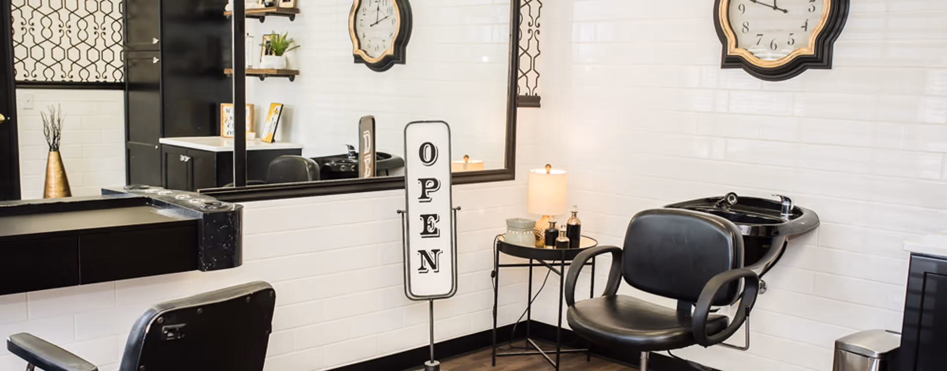 Interior of a salon area with two black salon chairs, a black hair washing sink, a large mirror, a small round table with a lamp and decorative items, a wall clock, and a vertical sign that reads OPEN. The walls are white tiled and the floor is dark wood.