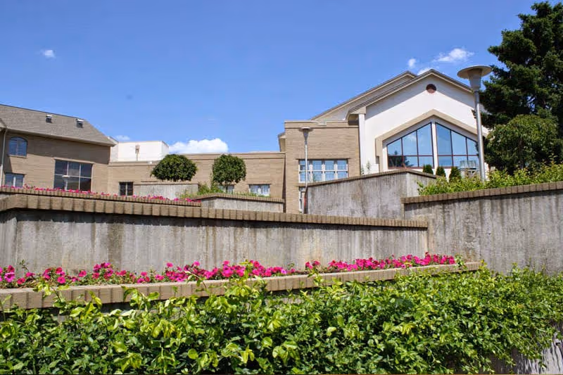 Exterior view of a senior living facility building with beige brick walls, large windows, and a landscaped area featuring green bushes and pink flowers under a clear blue sky.