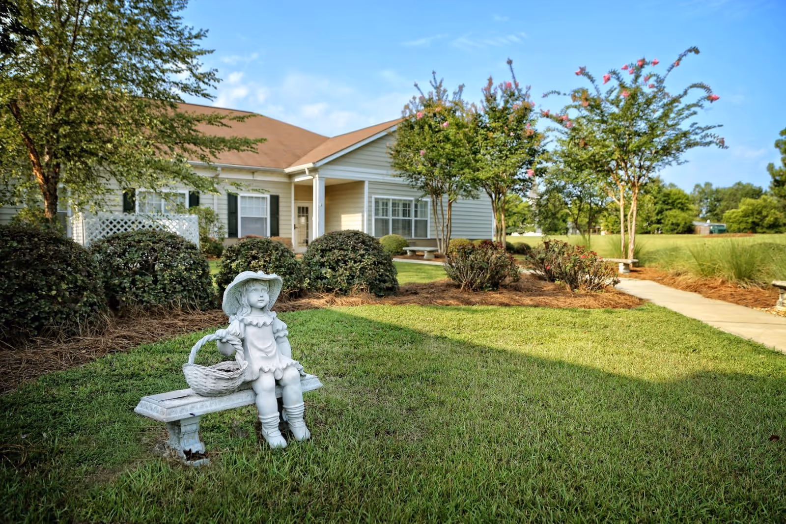 A small statue of a girl sitting on a bench on the lawn in front of a single-story senior care building with shrubs, trees, and a sidewalk.
