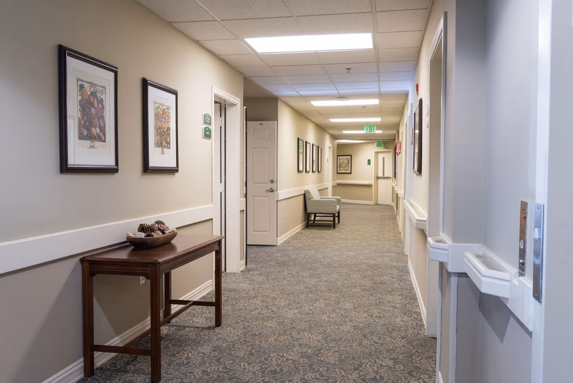A long, well-lit hallway in a senior living facility with beige walls and patterned carpet. The hallway features framed artwork on the walls, a wooden table with decorative pine cones, and a single upholstered chair along the wall. Several doors and exit signs are visible down the corridor.