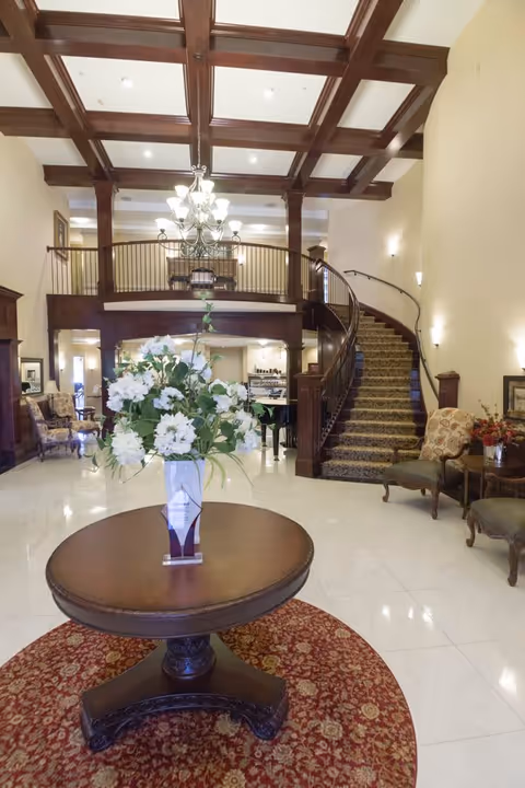 A spacious and elegant senior living facility lobby with a round wooden table in the center holding a vase of white flowers. The floor is tiled with a circular red patterned rug under the table. To the right, there are upholstered chairs and a side table with a flower arrangement. A curved staircase with a carpet runner and wooden railing leads to an upper level balcony with wooden beams on the ceiling and a chandelier hanging above.