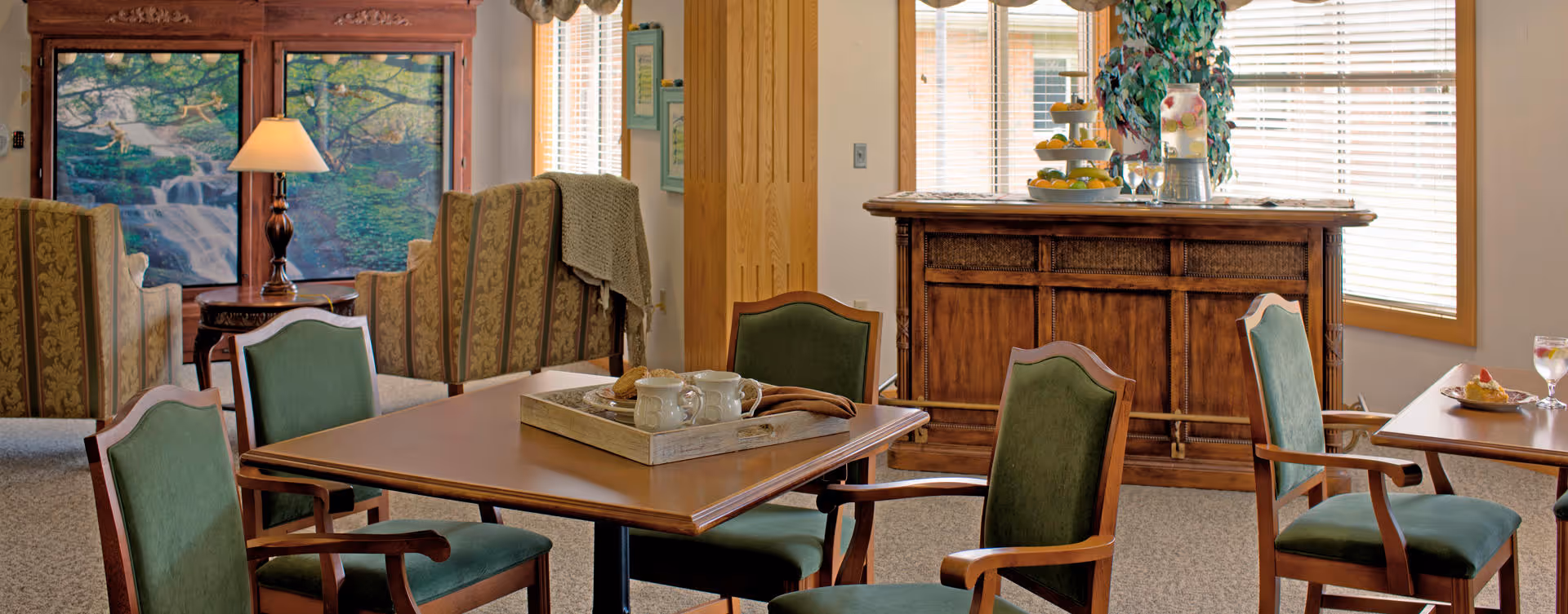 A cozy interior common area with wooden tables and green upholstered chairs. There is a wooden sideboard with decorative items and a large window with blinds letting in natural light. In the background, there are patterned armchairs and a table lamp in front of a large wall art depicting a waterfall scene.
