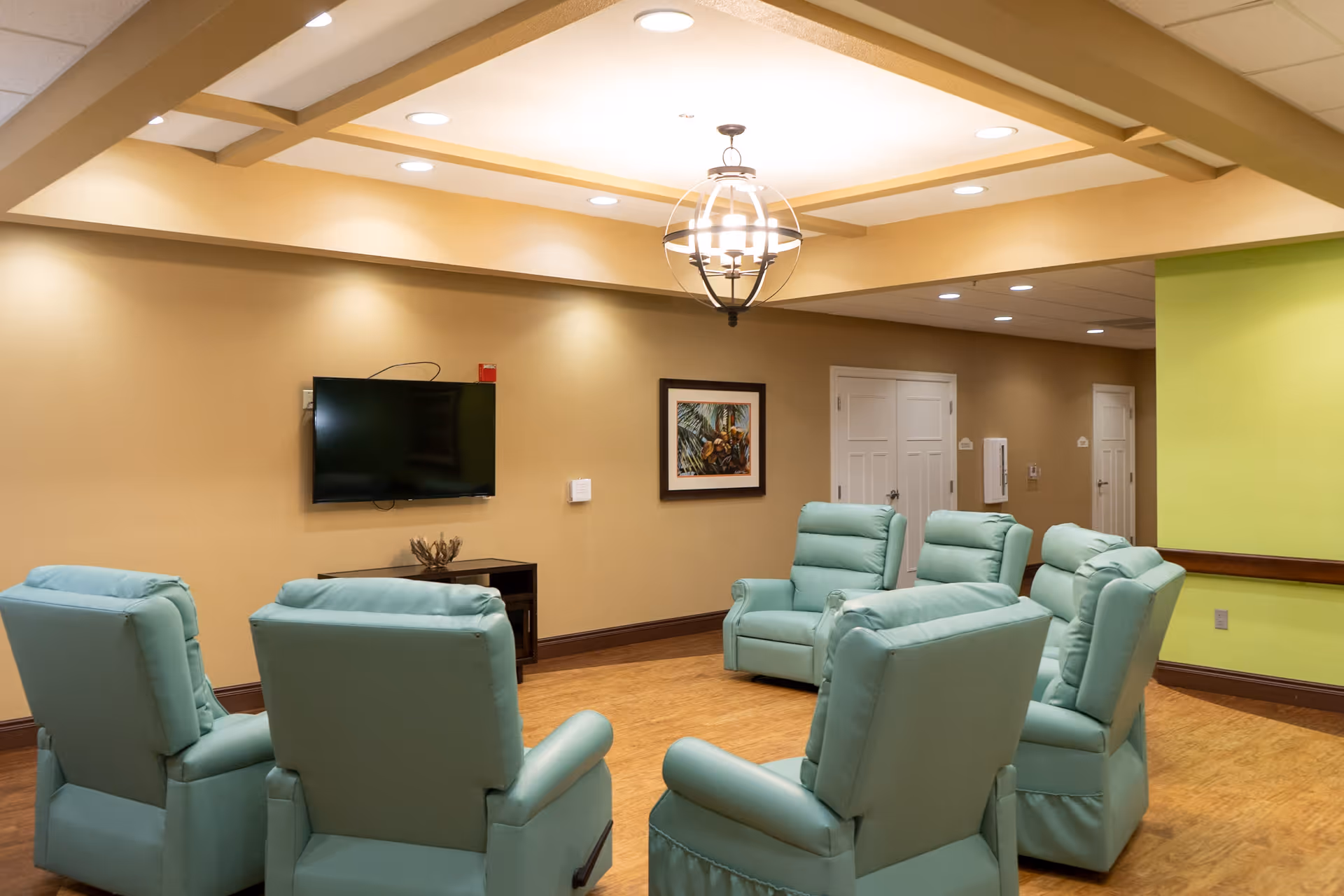 A cozy common area in an assisted living facility featuring six light blue recliner chairs arranged in a circle on a wooden floor. The walls are painted beige and light green, with a flat-screen TV mounted on one wall above a small cabinet. A framed picture hangs on the wall, and a modern chandelier light fixture is mounted on the ceiling.