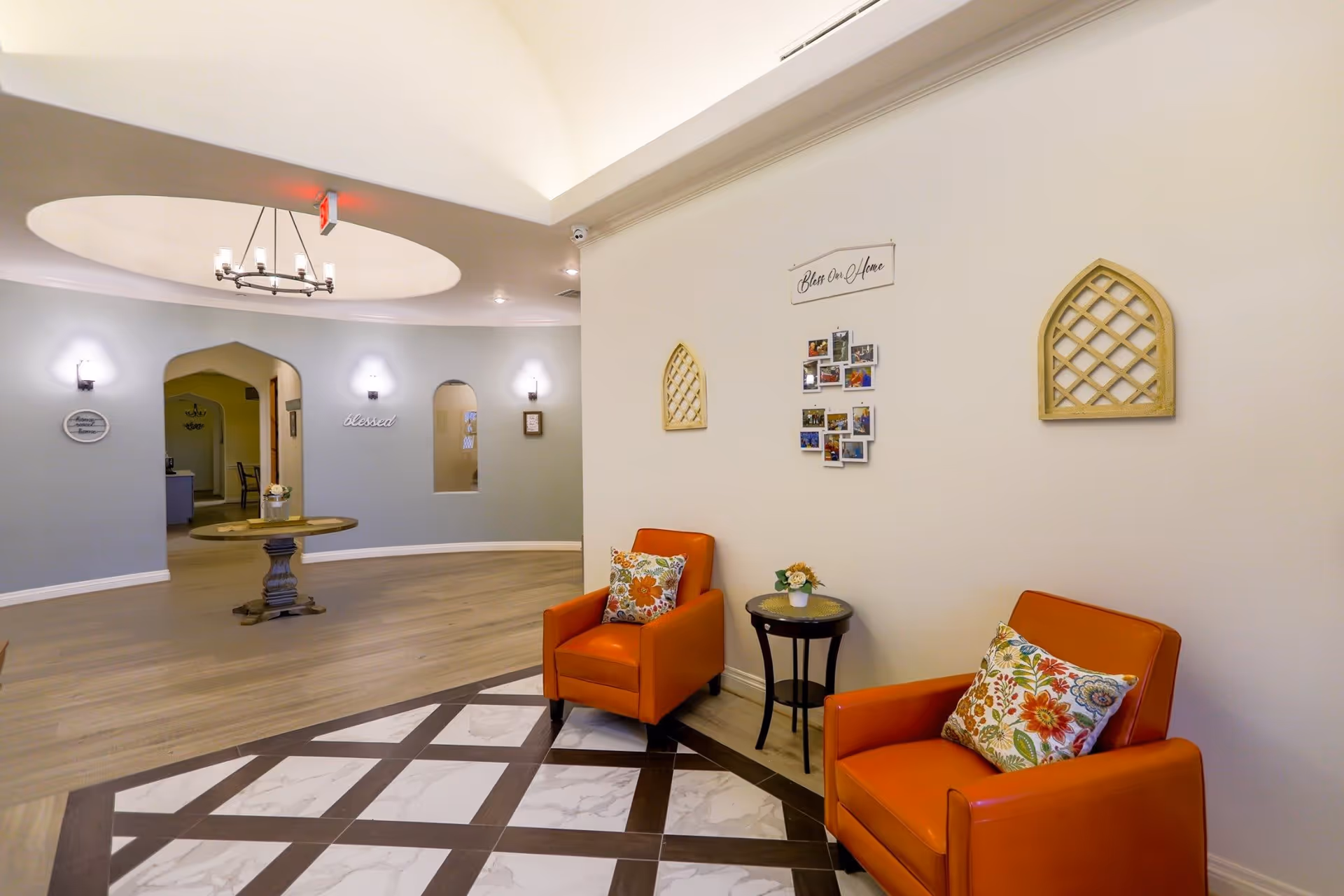 A cozy seating area in a memory care community with two orange armchairs adorned with floral cushions, a small round side table with a flower arrangement, and decorative wall hangings including a photo collage and lattice designs. The background shows a curved wall with arched doorways, soft lighting fixtures, and a round wooden table under a circular ceiling feature with a chandelier.