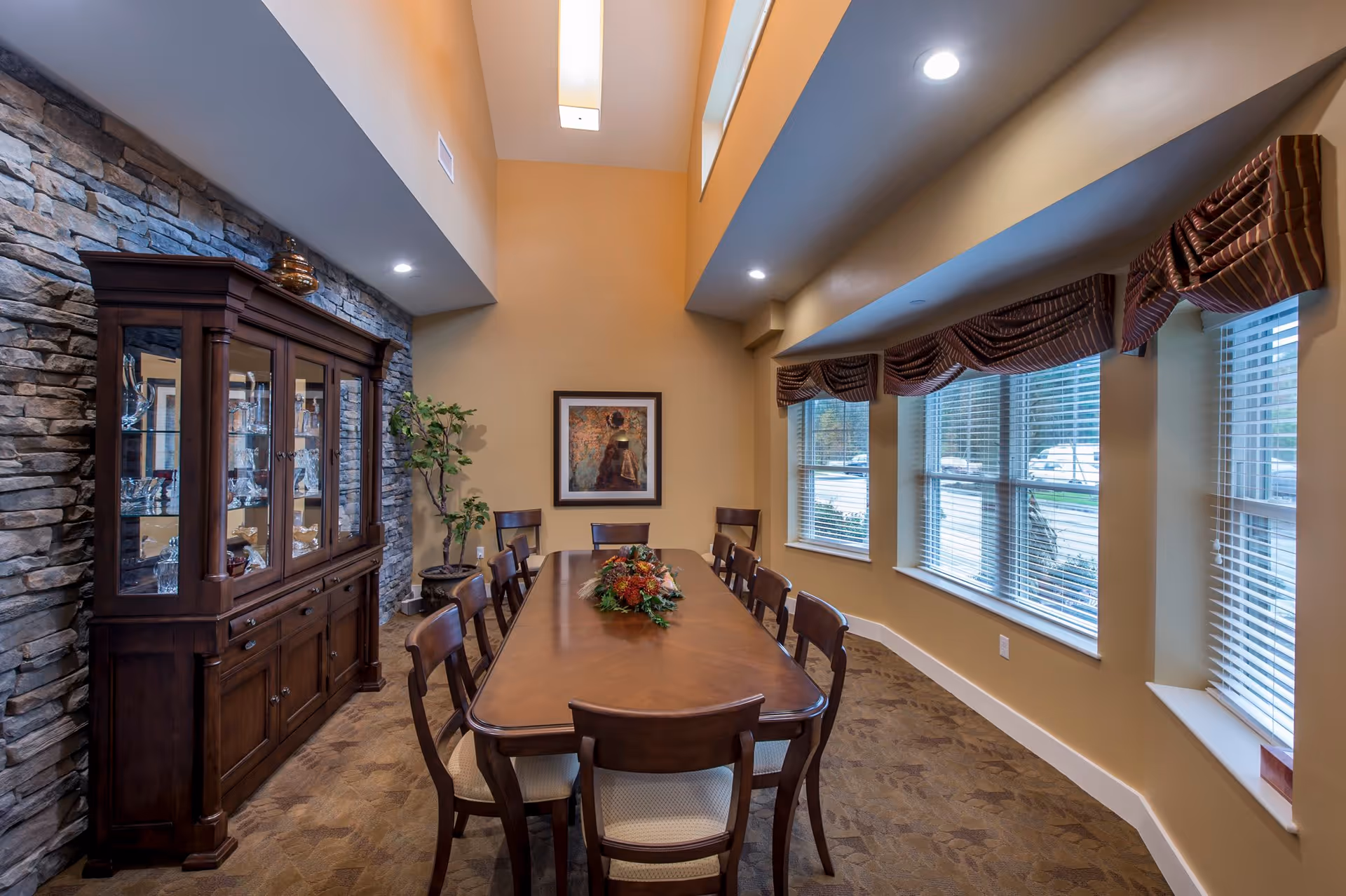 A dining room with a long wooden table surrounded by eight chairs. A floral centerpiece is placed on the table. To the left, there is a wooden china cabinet with glass doors displaying glassware. The wall behind the cabinet has a stone finish. On the right side, there are large windows with blinds and valances, allowing natural light into the room. The walls are painted beige, and the ceiling is high with recessed lighting.
