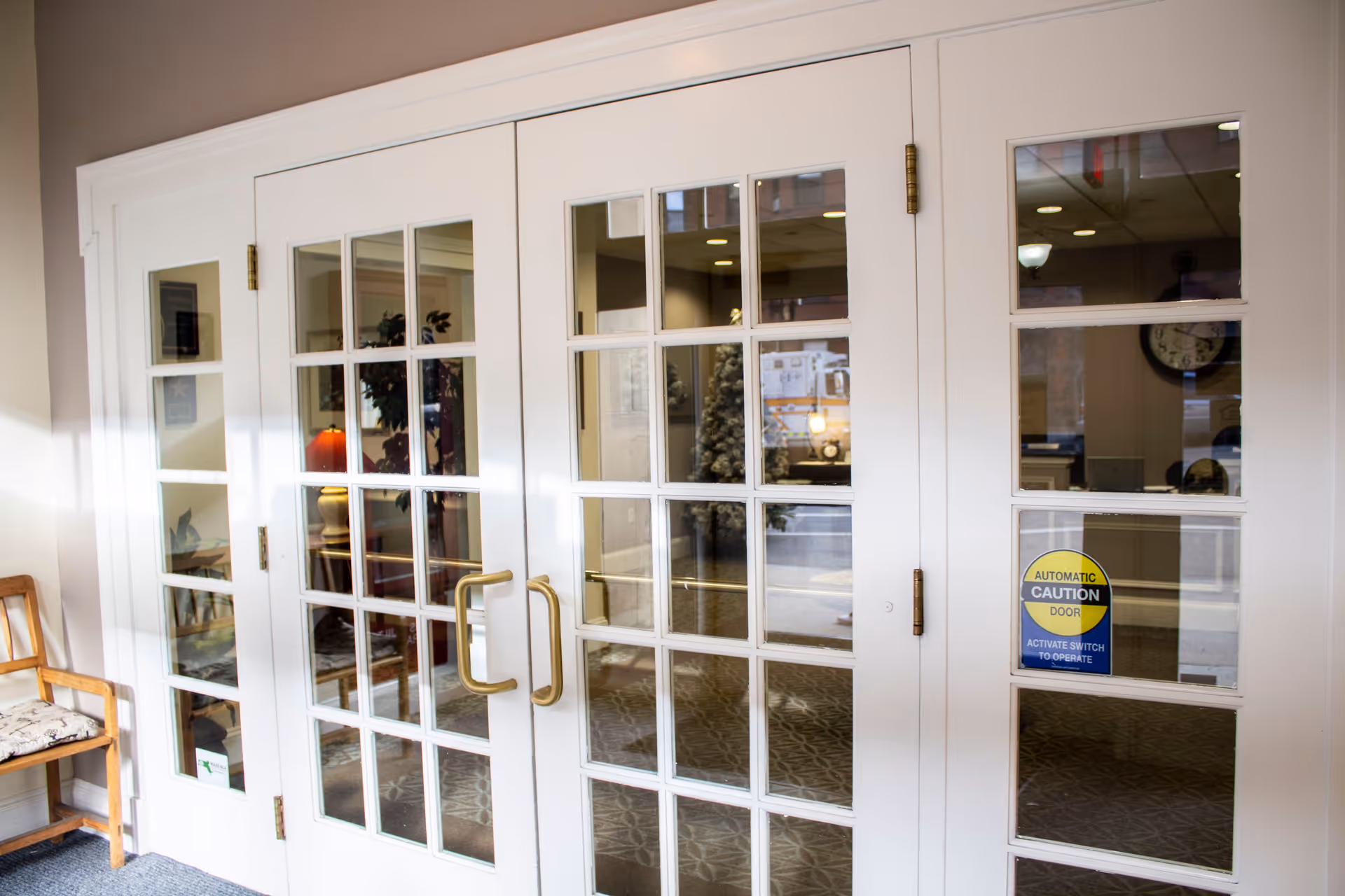 White double doors with multiple glass panes and brass handles leading into an interior space. A wooden chair with a patterned cushion is placed to the left of the doors. A sign on the right door reads 'Automatic Caution Door Activate Switch to Operate.' Inside, a clock and some furniture are visible through the glass panes.