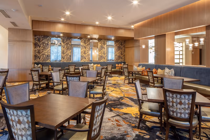 A spacious dining room with multiple wooden tables and cushioned chairs arranged neatly. The room features a patterned carpet with yellow and gray tones, a long cushioned bench along the wall with decorative pillows, and a wall with three vertical mirrors and floral wallpaper. The lighting is warm with recessed ceiling lights and modern hanging fixtures.