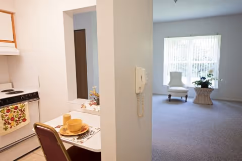 View of a small kitchen area with a stove, a table set with a cup and plate, and a chair. Adjacent to the kitchen is a living room with a large window, a white armchair, and a wicker side table with a plant on it.