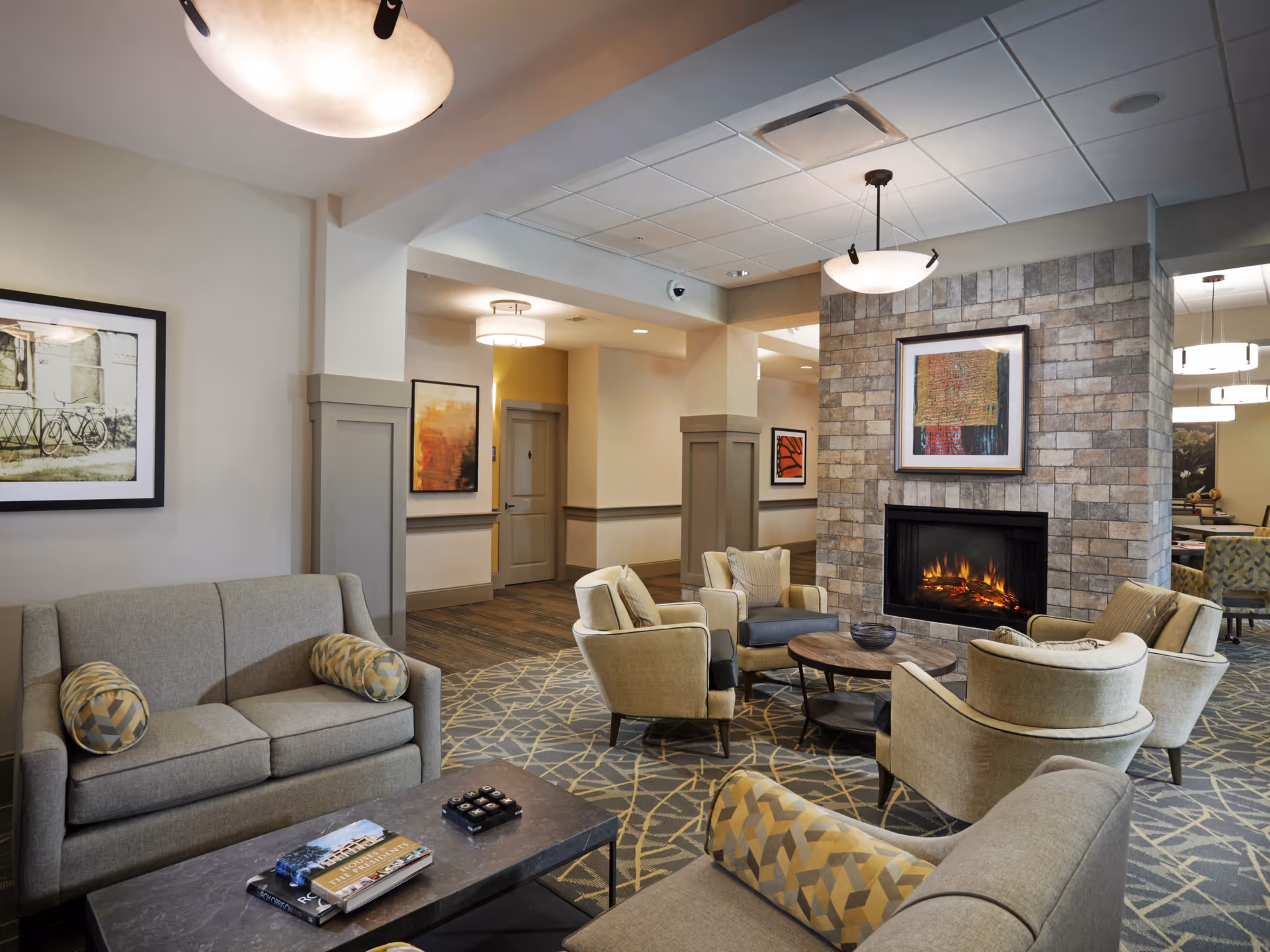 A cozy senior living facility common area with beige sofas and armchairs arranged around a round wooden coffee table in front of a stone fireplace with a framed artwork above it. The room features soft lighting from ceiling fixtures, patterned carpet, and framed pictures on the walls.