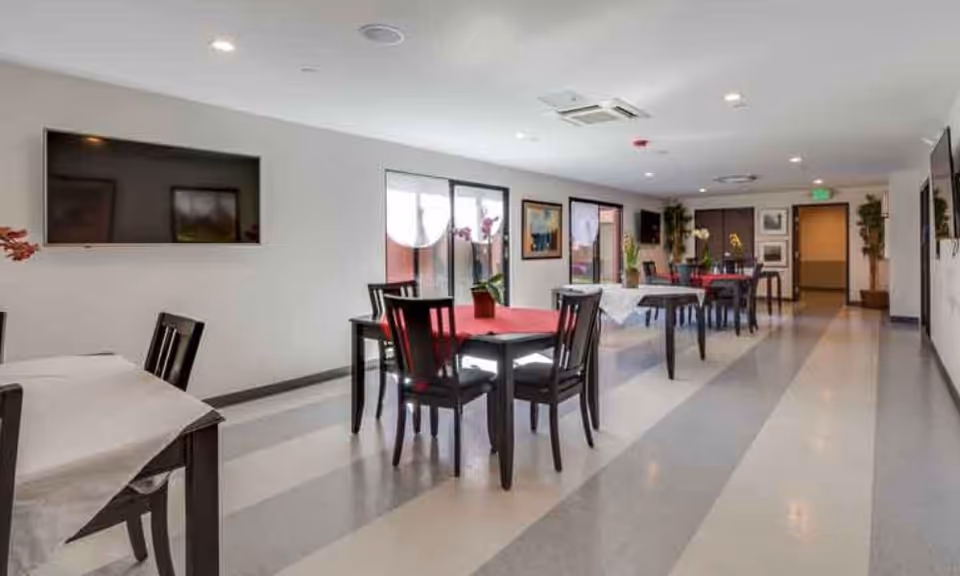 A bright dining room in a senior living facility with several tables covered with white and red tablecloths, each surrounded by black chairs. The room has large windows letting in natural light, a flat-screen TV mounted on the wall, and framed artwork. There are plants and decorative items placed around the room.