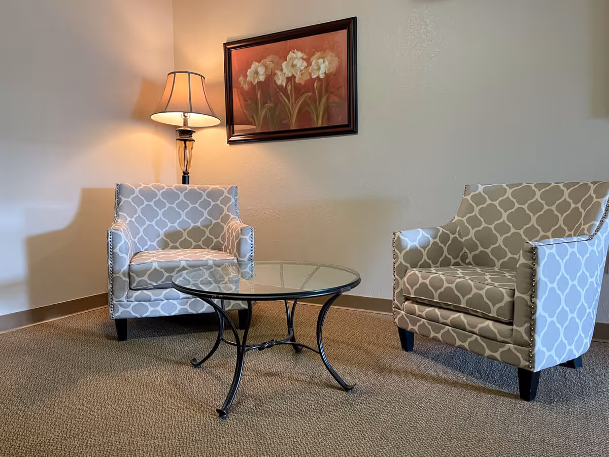 Two patterned armchairs with a glass-top round metal table between them, a lit table lamp behind one chair, and a framed painting of white flowers on the wall in a cozy room.