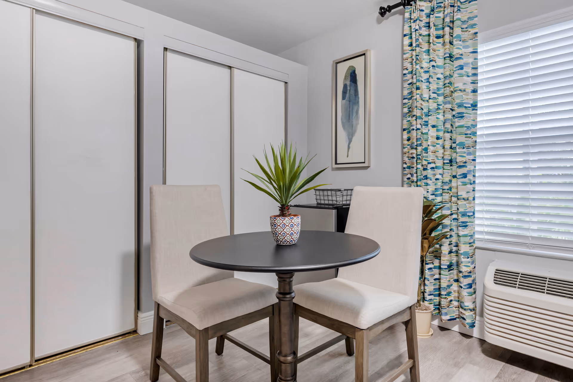 Small dining area with a round table, two upholstered chairs, a potted plant centerpiece, and a window with blinds and patterned curtains.