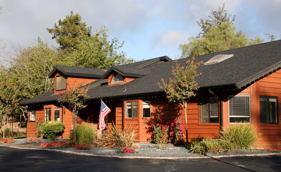 Single-story red wooden building with landscaped front, an American flag, and a dark roof under a partly cloudy sky.