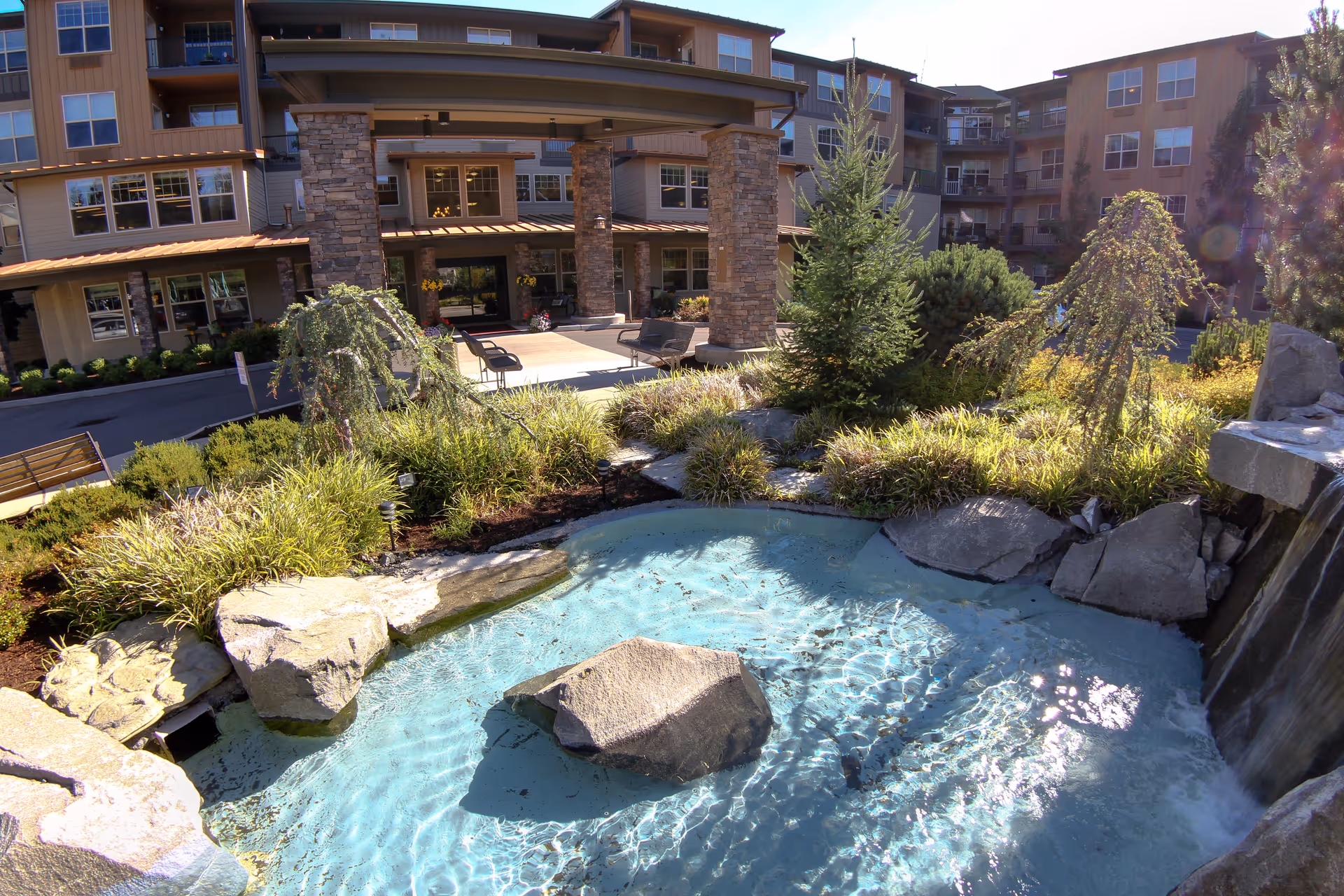 Outdoor view of a senior living facility with a landscaped garden featuring a small pond with clear water and rocks, surrounded by greenery and trees. The building in the background has multiple floors with balconies and large windows, and a covered entrance supported by stone pillars.
