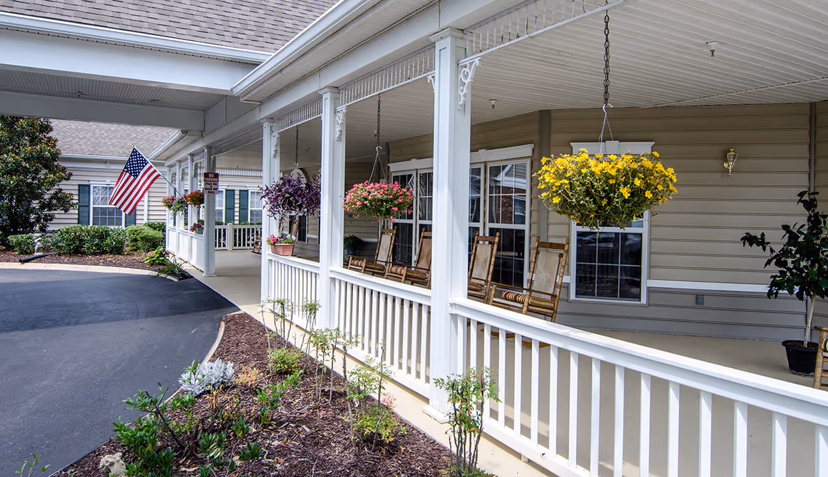 Covered front porch of a senior living facility with rocking chairs, hanging flower baskets, and an American flag.