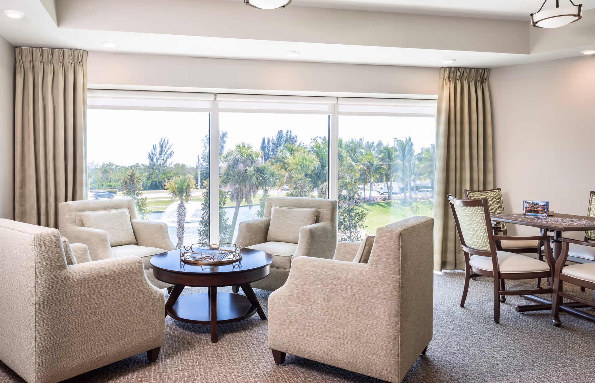 A bright and spacious living room area with four beige armchairs arranged around a round wooden coffee table. Large windows with beige curtains offer a view of palm trees and greenery outside. To the right, there is a dining table with four chairs featuring patterned upholstery.