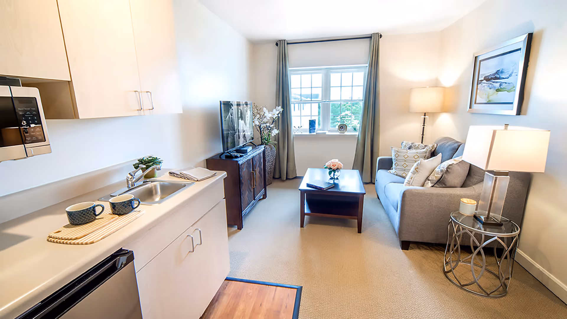 A bright and cozy living area in a senior living facility featuring a small kitchenette with a sink, microwave, and two cups on a tray. The room has a gray sofa with decorative pillows, a wooden coffee table with a small flower vase, a side table with a lamp, a floor lamp, a TV on a wooden stand, and a window with curtains letting in natural light.