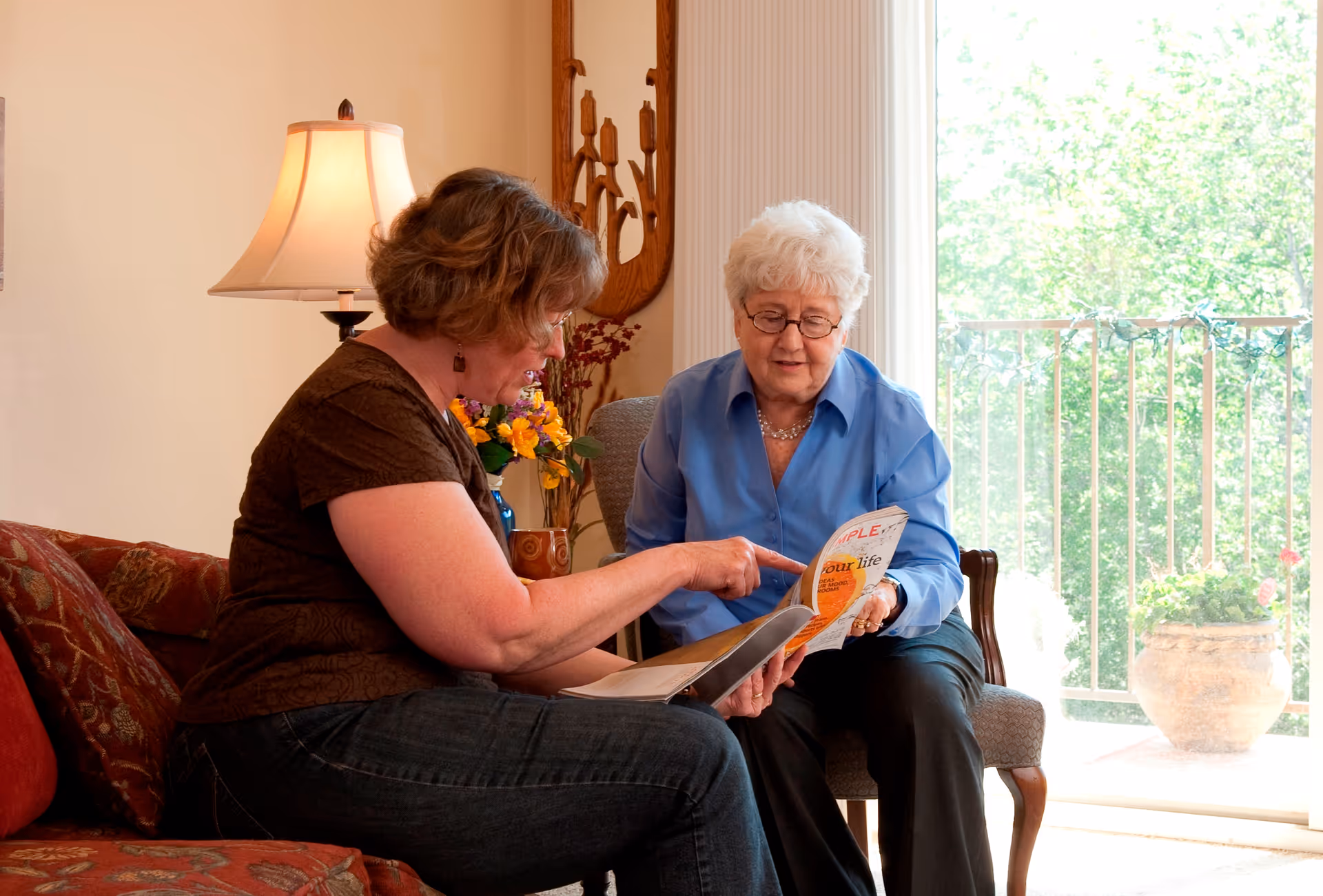 Two women sitting in a living room area, one older woman with white hair and glasses wearing a blue shirt, and a younger woman with brown hair wearing a brown top and jeans. They are looking at and discussing a magazine or booklet together. The room has a lamp, floral arrangement, and a large window with a view of greenery outside.