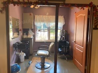 Small salon room with a styling chair, mirror, hair tools on a cart, and a window with curtains.