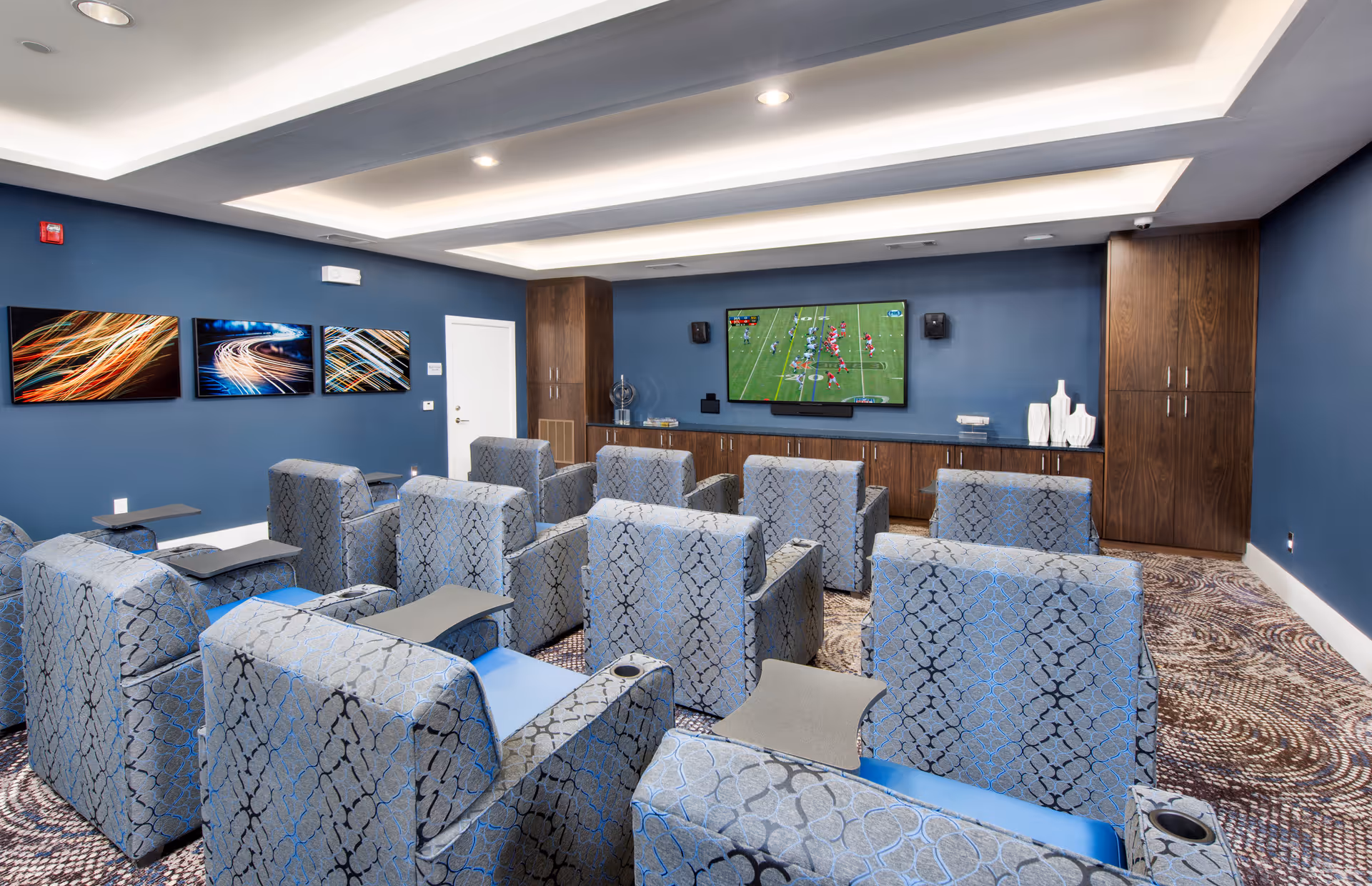 A small theater-style media room with upholstered recliner seats arranged in rows facing a wall-mounted TV showing a football game.