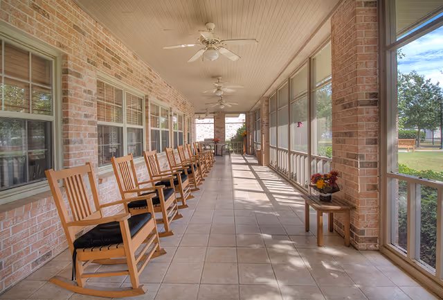 A long, covered outdoor porch area with a row of wooden rocking chairs lined up against a brick wall with windows. The porch has ceiling fans and tiled flooring, with a small table holding a flower arrangement near the screened side. Outside, there is greenery and a bench visible through the screened windows.