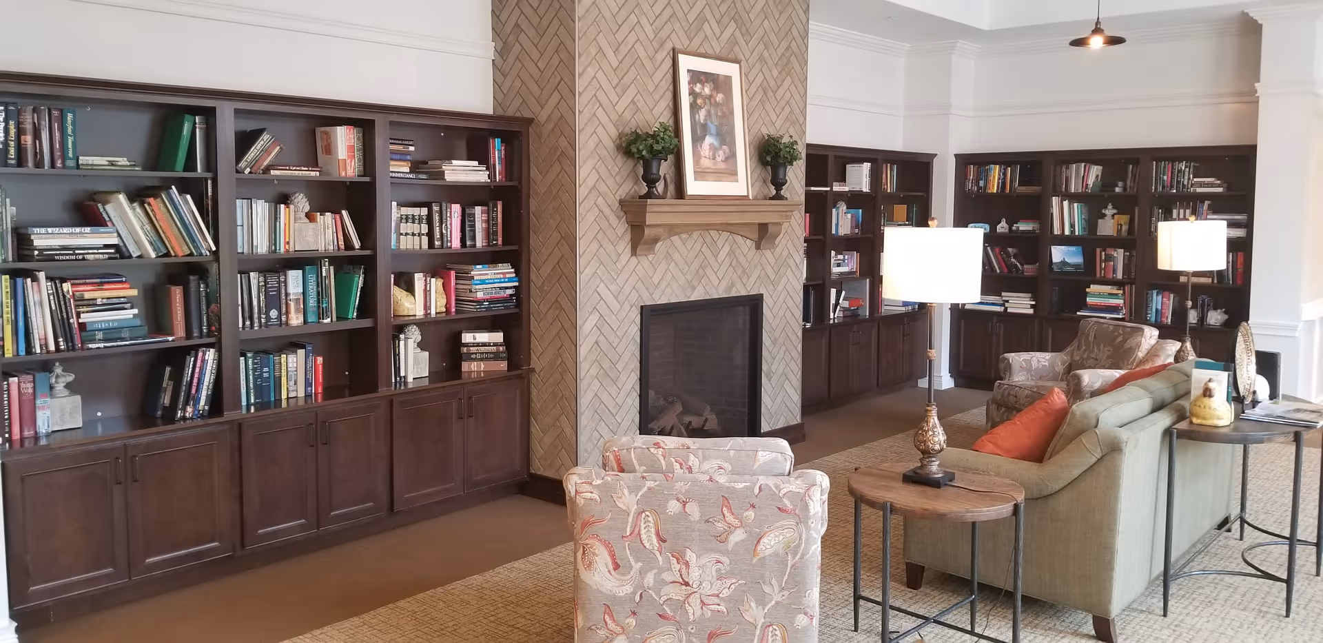 A cozy living room area with built-in dark wood bookshelves filled with books, a fireplace with a herringbone-patterned tile surround, and comfortable seating including a green sofa with orange pillows and two patterned armchairs. There are two table lamps on wooden side tables, and a framed floral picture above the fireplace.