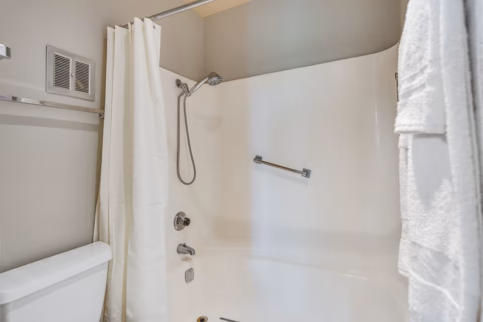 A clean bathroom featuring a white bathtub with a showerhead and a white shower curtain. There is a towel rack with white towels hanging on the right side and a toilet partially visible on the left side.