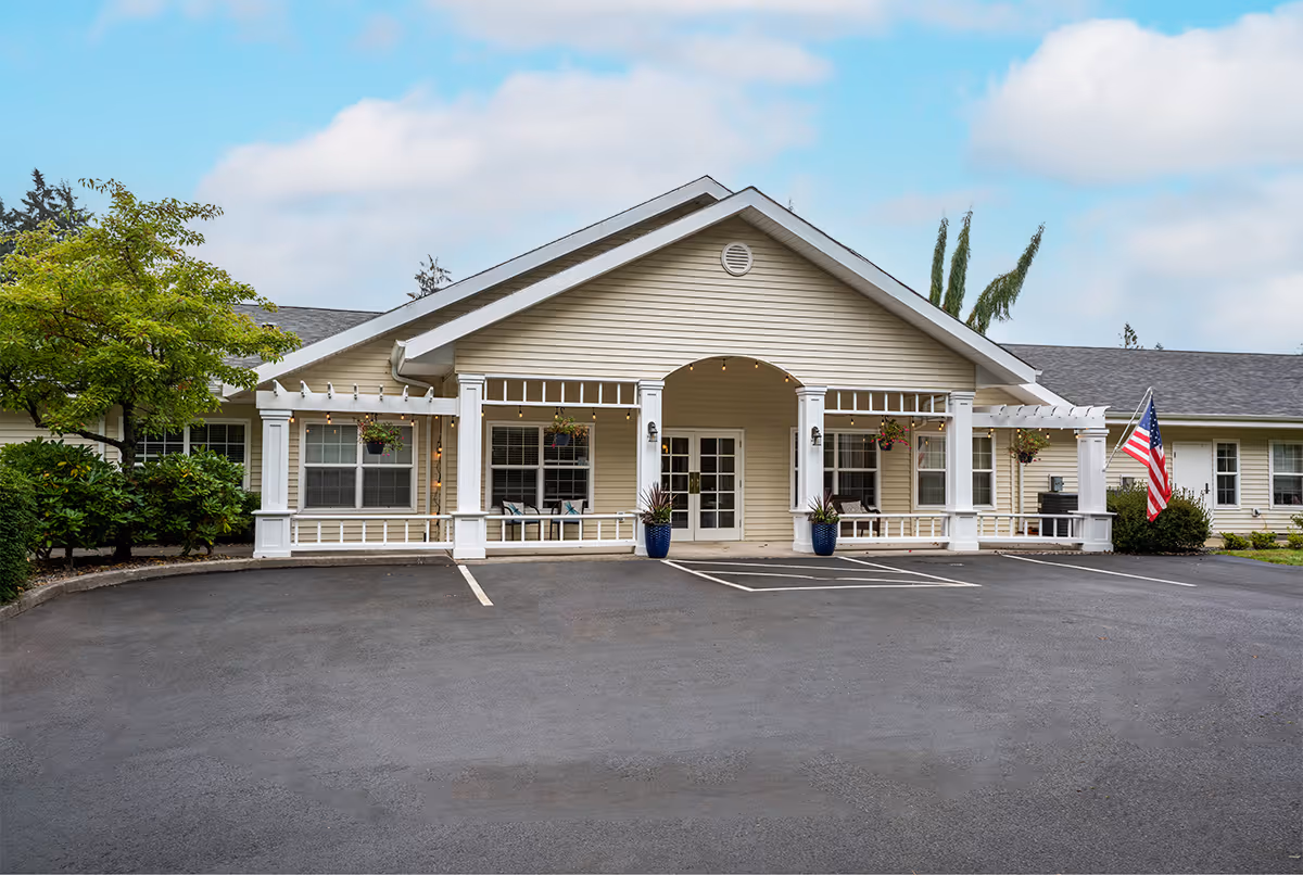 Front exterior view of a single-story building with beige siding and white trim. The entrance features a covered porch with columns, hanging flower pots, and string lights. There are two large blue planters with plants near the entrance doors. An American flag is displayed on the right side near some bushes. The parking area in front is empty with marked parking spaces.