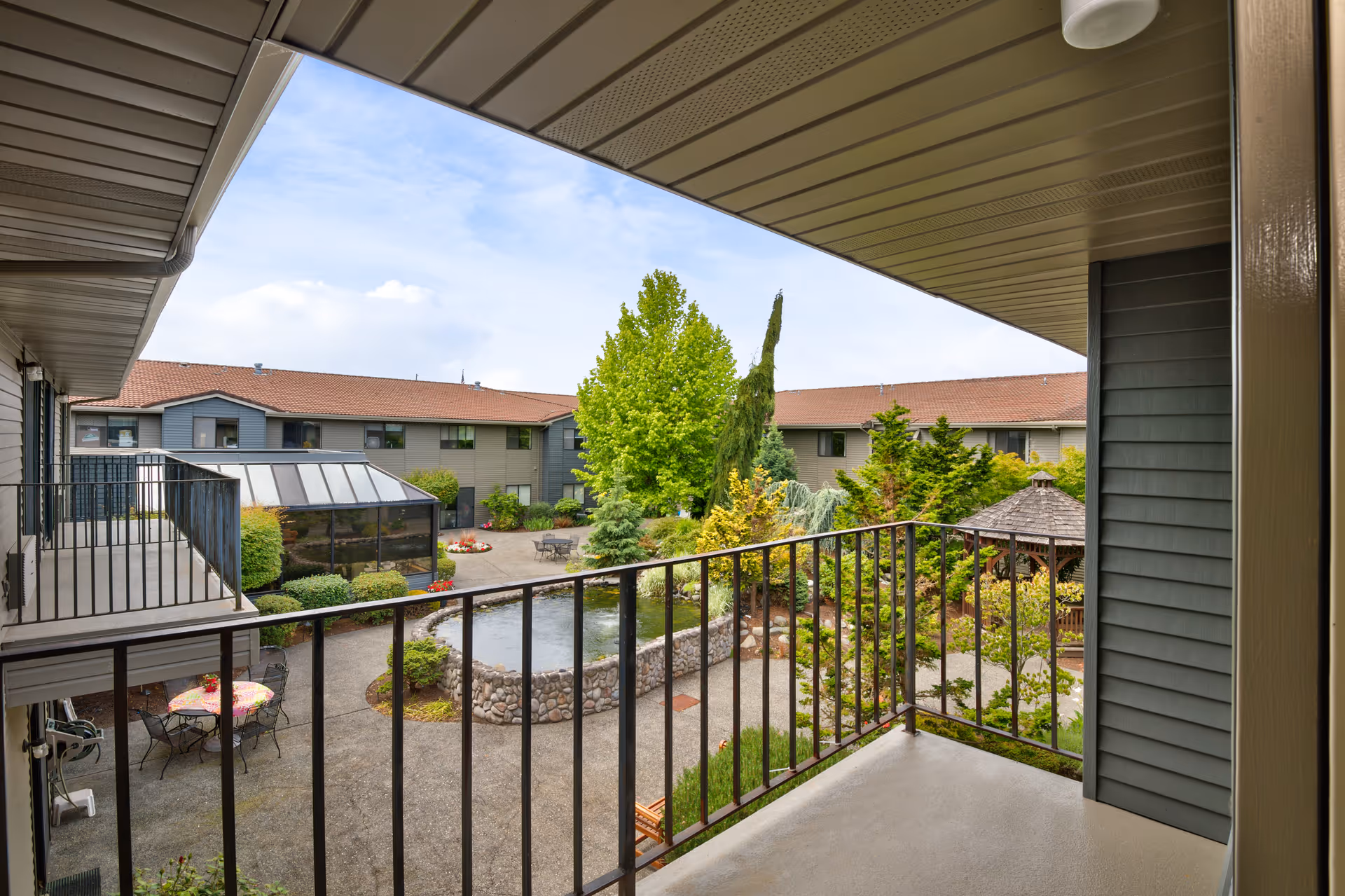View from a covered balcony overlooking a landscaped courtyard with a stone-bordered pond, various trees and shrubs, outdoor seating areas with tables and chairs, and a gazebo. The courtyard is surrounded by a two-story building with a red-tiled roof and gray siding.