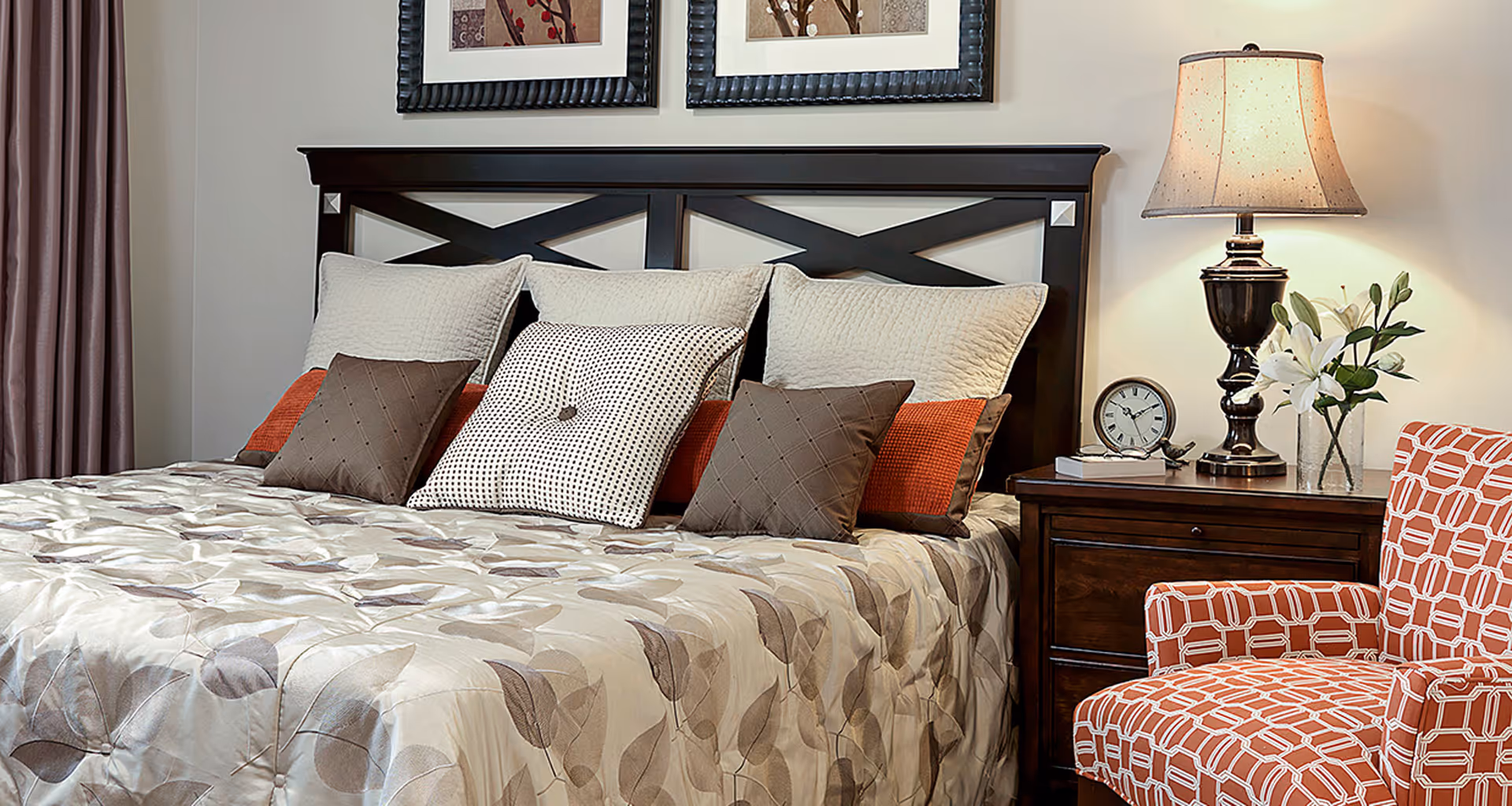 Well-decorated bedroom with a large bed topped with multiple pillows, a bedside table with a lamp and clock, and an upholstered chair.