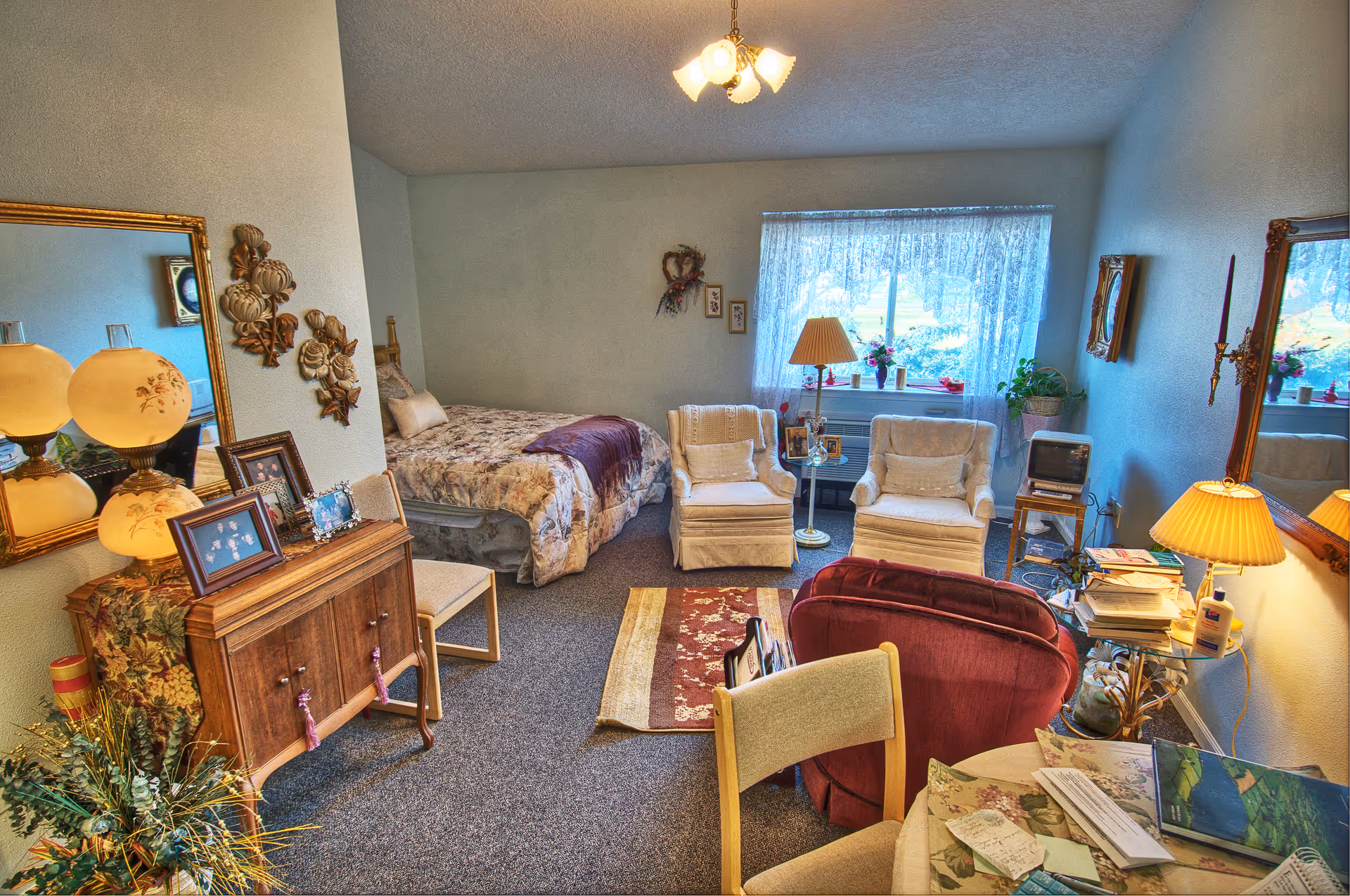 A cozy assisted living room with a bed covered in floral bedding, two armchairs near a window with lace curtains, a small table with a lamp, a wooden cabinet with framed photos, a red armchair, and a round table with papers and a lamp. The room is warmly lit and decorated with plants and wall art.