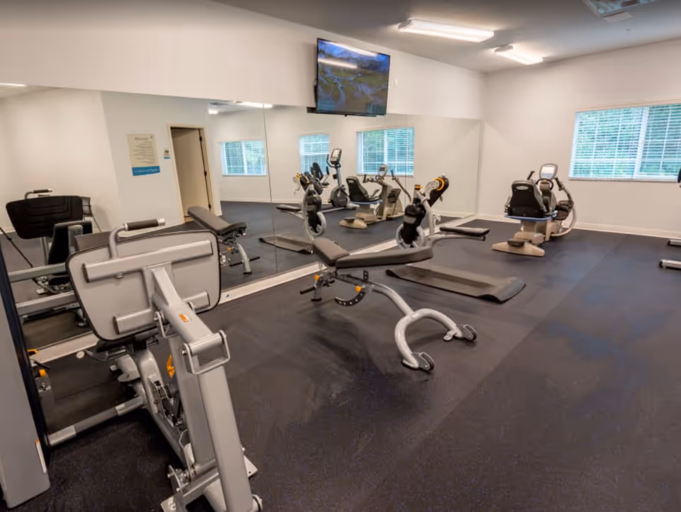 Interior view of a fitness room with exercise equipment including a weight machine, a bench, recumbent bikes, and a treadmill. The room has large mirrors on one wall, a window with blinds, and a wall-mounted TV.