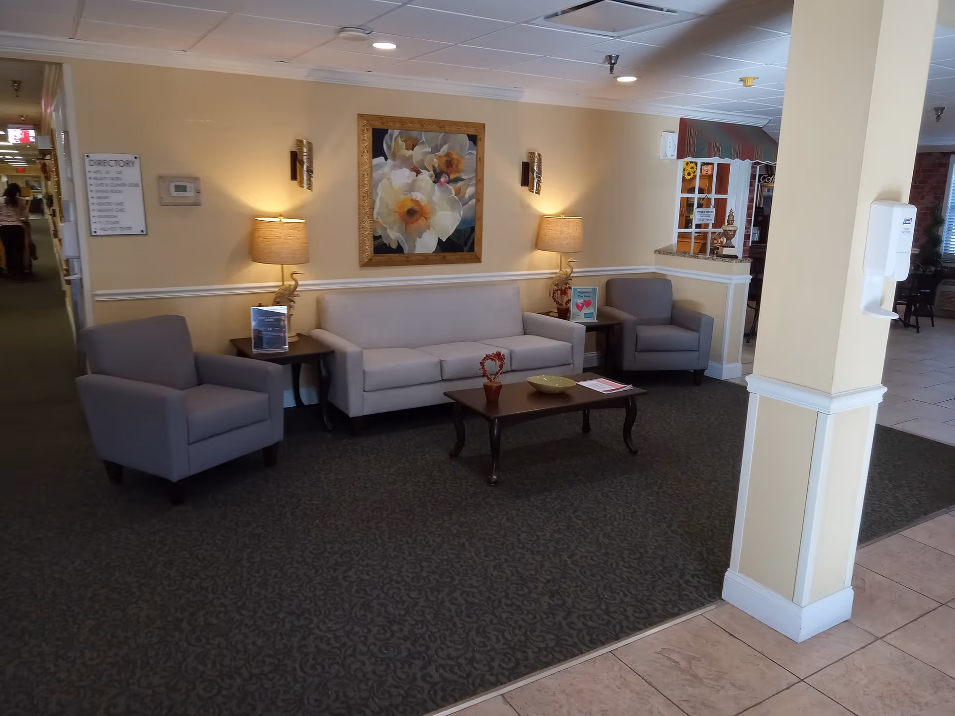 A cozy seating area in a senior living facility with a beige sofa, two gray armchairs, a wooden coffee table with decorative items, two table lamps with beige shades, and a large floral painting on the wall. The area has carpeted flooring and is adjacent to a hallway and a tiled floor section.