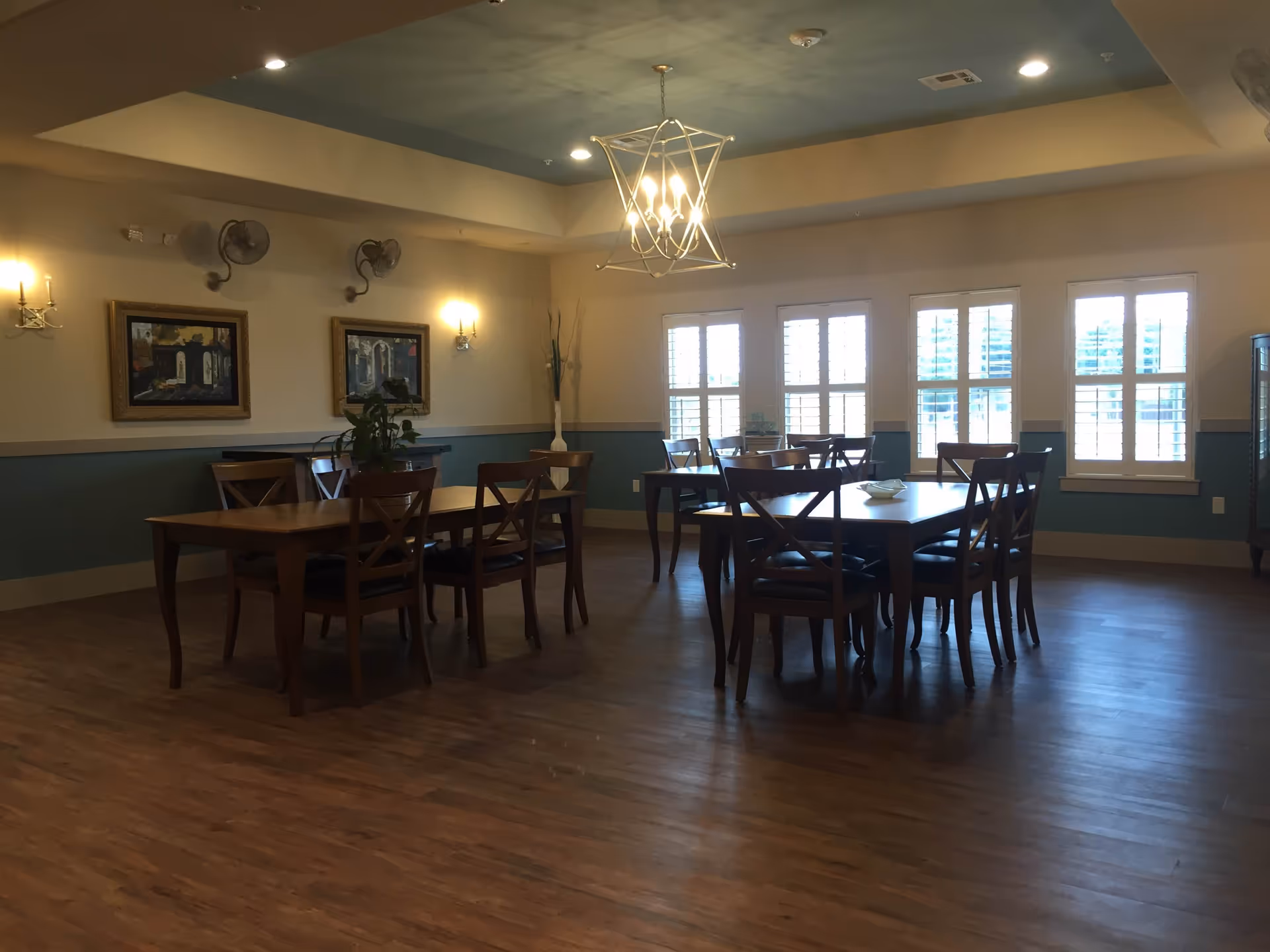 A dining room with wooden tables and chairs arranged neatly. The room has light-colored walls with a blue accent wall and wooden flooring. There are three windows with white shutters letting in natural light. Two framed paintings hang on the wall, and a modern chandelier hangs from the ceiling. Wall-mounted fans and sconces provide additional lighting.