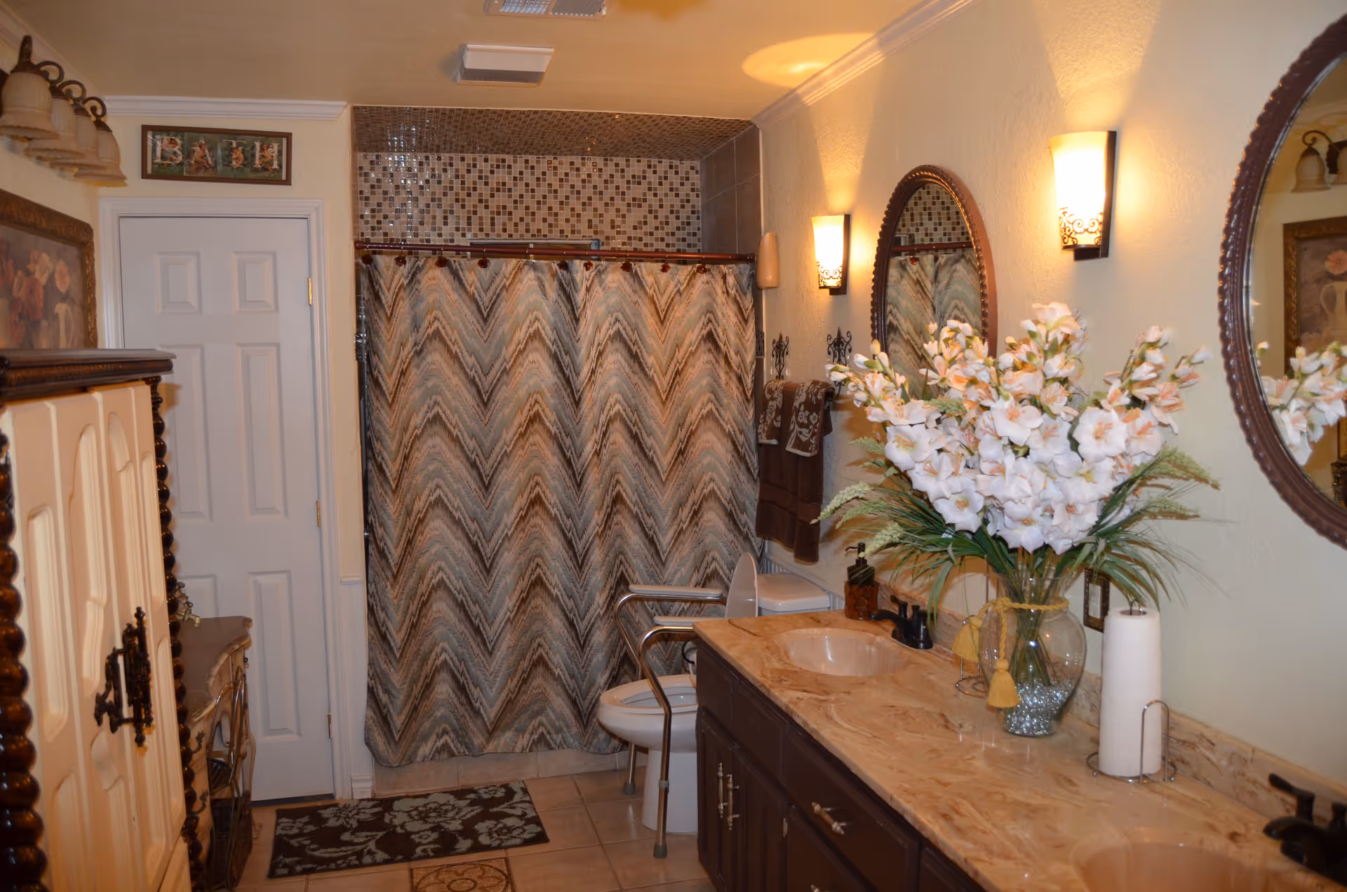 A bathroom with a double sink vanity topped with a marble countertop, a large vase of white flowers, and a paper towel holder. There are two oval mirrors above the sinks with wall-mounted light fixtures on either side. A toilet with a safety frame is next to a shower with a patterned curtain and mosaic tile accent. A white door is visible on the left side with a framed sign above it that reads 'BATH'. The floor has patterned rugs and there is a wooden cabinet on the left side.