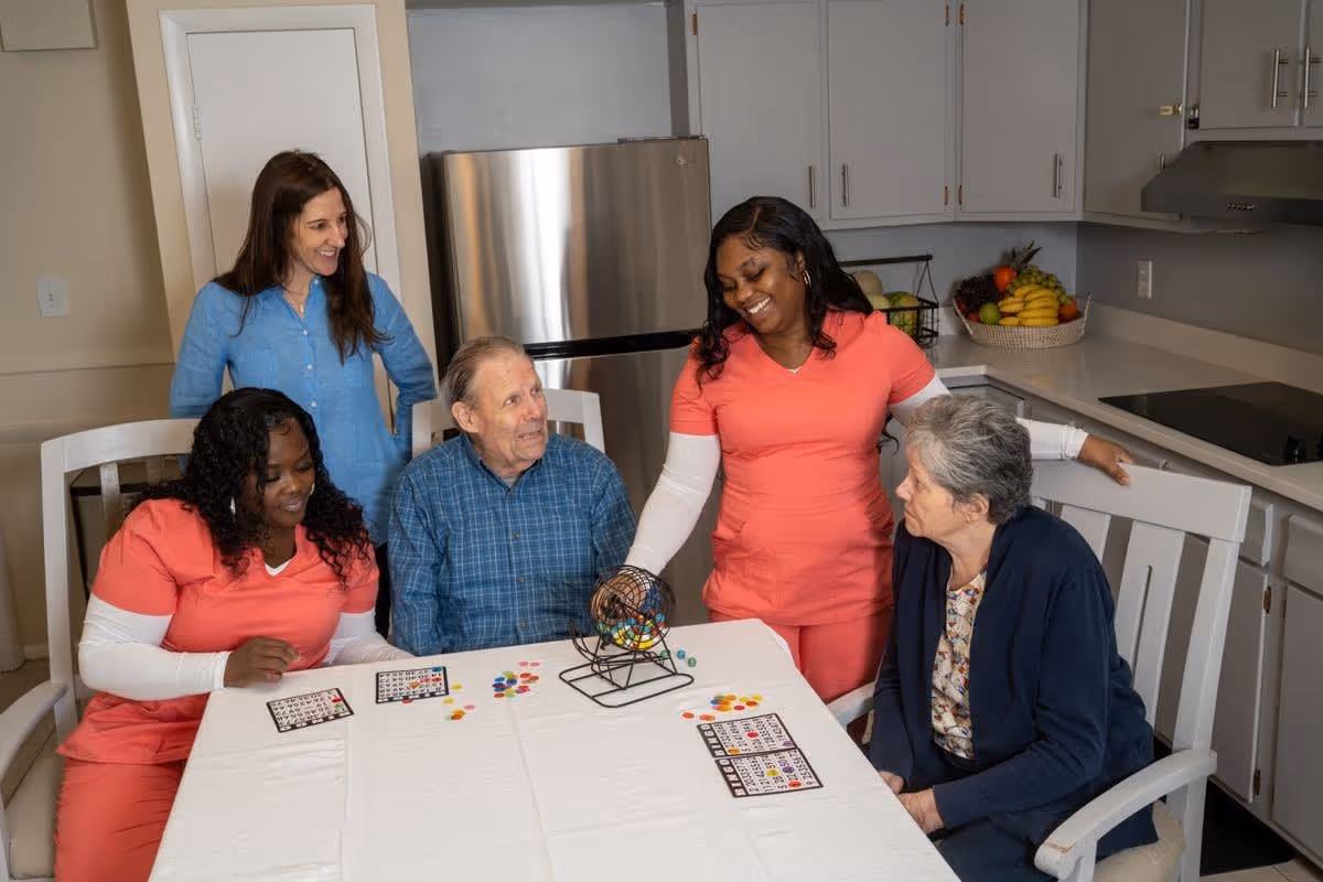 Two elderly individuals and three caregivers playing bingo together at a table in a kitchen area. The caregivers are wearing coral scrubs and one woman in a blue shirt is standing behind them, smiling. The kitchen has white cabinets, a stainless steel refrigerator, and a basket of fruit on the counter.
