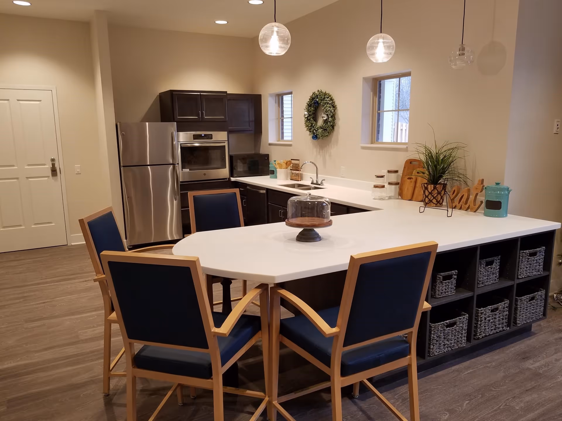 Modern kitchen area with a large white island countertop surrounded by four wooden chairs with blue cushions. The kitchen features dark cabinets, a stainless steel refrigerator, built-in oven, microwave, and a sink under two small windows. Decorative items include a wreath on the wall, a plant, and kitchen containers on the counter.