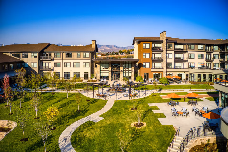 Aerial view of a multi-story senior living building overlooking a landscaped courtyard with winding paths, seating areas, and orange umbrellas.